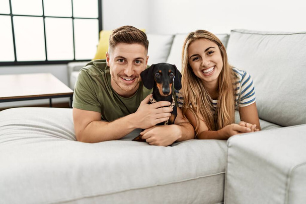 Young hispanic couple smiling happy lying on the sofa with dog at home. - 写真・画像