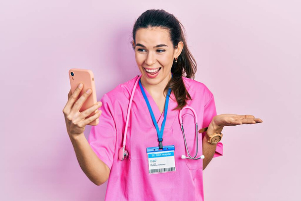 Mladá brunetka lékař žena dělá videohovor konzultace s chytrým telefonem slaví úspěch se šťastným úsměvem a vítězný výraz se zvedl ruku  - Fotografie, Obrázek