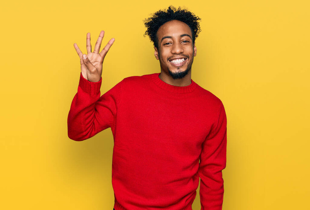 Νεαρός Αφροαμερικάνος με γενειάδα που φοράει χειμωνιάτικο πουλόβερ που δείχνει και δείχνει με τα δάχτυλα νούμερο τέσσερα ενώ χαμογελά σίγουρος και χαρούμενος.  - Φωτογραφία, εικόνα