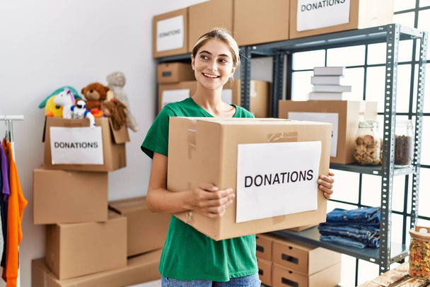 Joven mujer caucásica vistiendo uniforme voluntario sosteniendo caja de donaciones en el centro de caridad - Foto, imagen