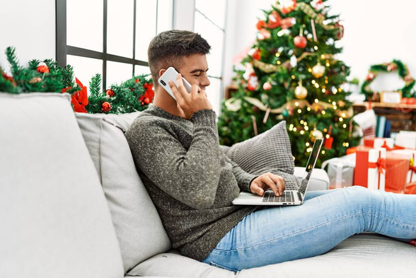 Молодой латиноамериканец использует ноутбук и разговаривает на смартфоне сидя на диване у рождественской елки дома - Фото, изображение