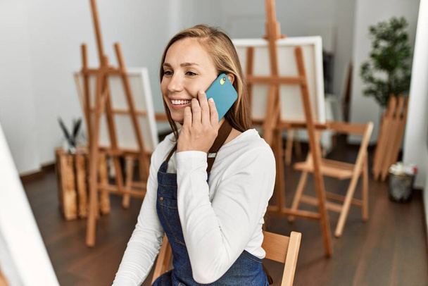 Mladá běloška žena usmívá sebevědomě mluvit na smartphone v ateliéru umění - Fotografie, Obrázek