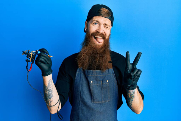 Redhead άνθρωπος με μακριά γενειάδα καλλιτέχνης τατουάζ φορώντας επαγγελματική στολή και γάντια χαμογελώντας με χαρούμενο πρόσωπο κλείσιμο του ματιού στην κάμερα κάνει σημάδι νίκη με τα δάχτυλα. Νούμερο δύο..  - Φωτογραφία, εικόνα