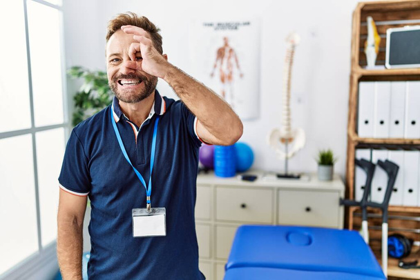 Hombre fisioterapeuta de mediana edad que trabaja en la clínica de recuperación del dolor haciendo buen gesto con la mano sonriente, los ojos mirando a través de los dedos con la cara feliz.  - Foto, Imagen