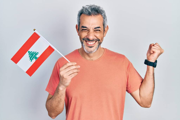 Όμορφος μεσήλικας άντρας με γκρίζα μαλλιά που κρατάει τη σημαία του Λιβάνου ουρλιάζοντας περήφανος, γιορτάζοντας τη νίκη και την επιτυχία πολύ ενθουσιασμένος με το σηκωμένο χέρι  - Φωτογραφία, εικόνα
