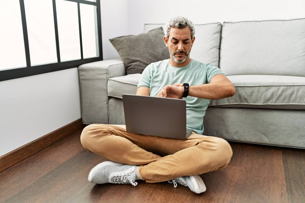 Middelbare leeftijd Spaanse man met behulp van laptop zitten op de vloer in de woonkamer controleren van de tijd op de pols horloge, ontspannen en vol vertrouwen  - Foto, afbeelding