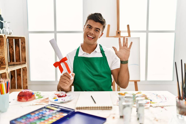 Young hispanic man at art studio holding degree waiving saying hello happy and smiling, friendly welcome gesture  - Photo, Image