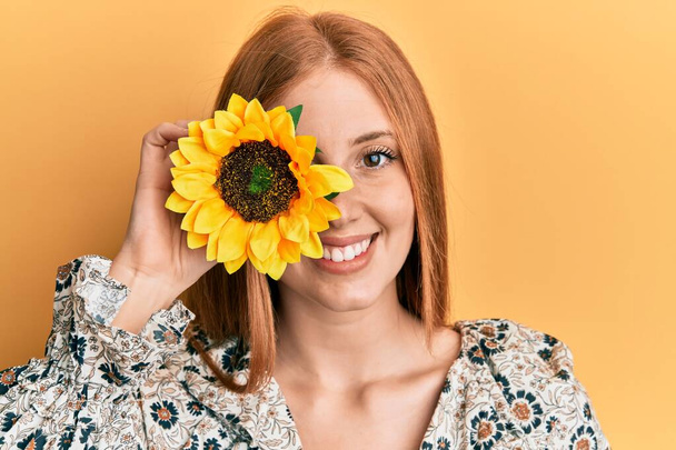 Young irish woman holding sunflower over eye looking positive and happy standing and smiling with a confident smile showing teeth  - Fotoğraf, Görsel