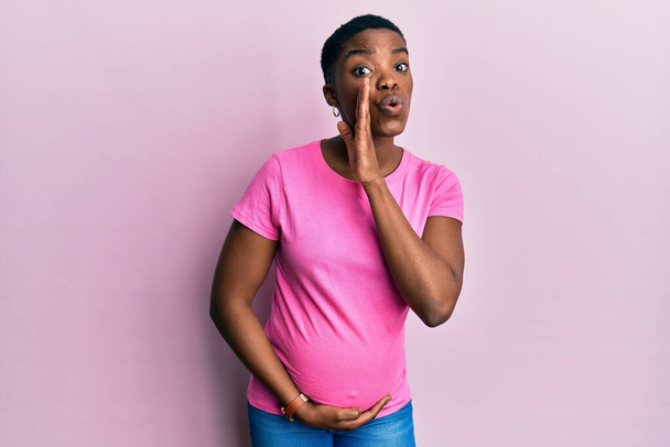 Young african american woman expecting a baby, touching pregnant belly hand on mouth telling secret rumor, whispering malicious talk conversation  - Photo, Image