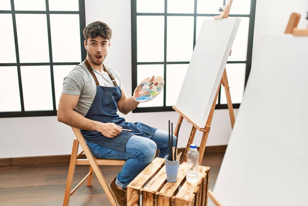 Молодой латиноамериканец рисует в художественной студии испуганный и изумленный с открытым ртом в ожидании сюрприза, недоверия  - Фото, изображение