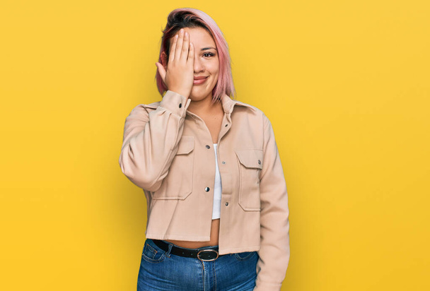 Hispanic woman with pink hair wearing casual clothes covering one eye with hand, confident smile on face and surprise emotion.  - Photo, Image