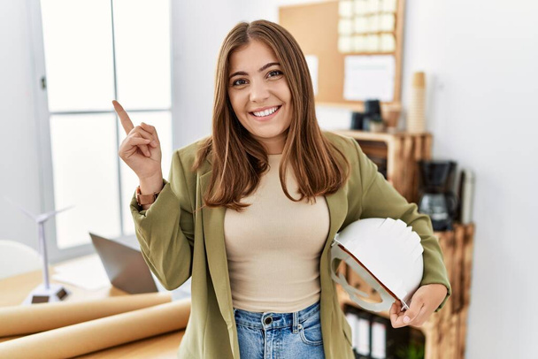 Junge brünette Frau mit Architektenhut im Büro lächelt glücklich und zeigt mit Hand und Finger zur Seite  - Foto, Bild