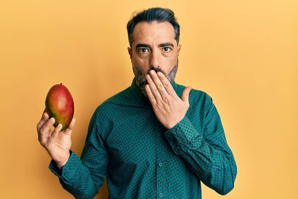 Мужчина средних лет с бородой и седыми волосами держит манго, прикрывая рот рукой, шокирован и боится ошибиться. Удивительное выражение  - Фото, изображение