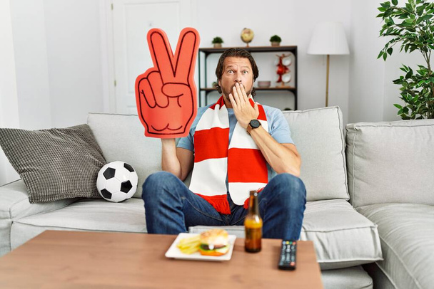 Guapo hombre de mediana edad hooligan fútbol animando juego en casa cubriendo la boca con la mano, sorprendido y temeroso de error. expresión sorprendida  - Foto, imagen