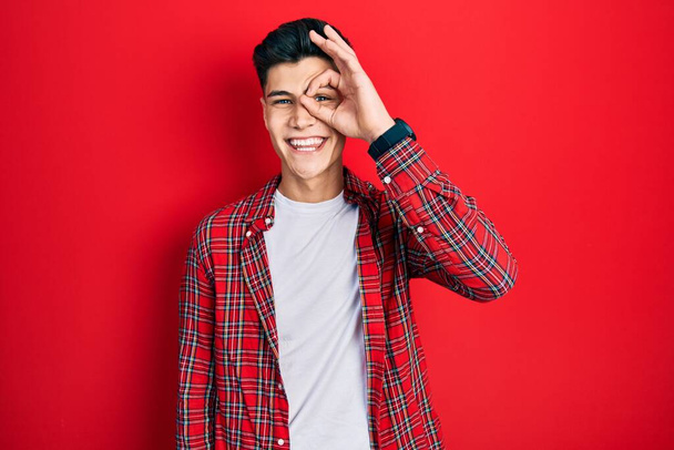 Jonge Latijns-Amerikaanse man draagt casual kleding doet ok gebaar met de hand glimlachen, oog kijken door vingers met gelukkig gezicht.  - Foto, afbeelding