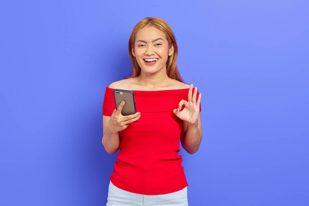 Portrait of smiling young Asian woman holding mobile phone and gesturing okay sign isolated on purple background - Fotoğraf, Görsel