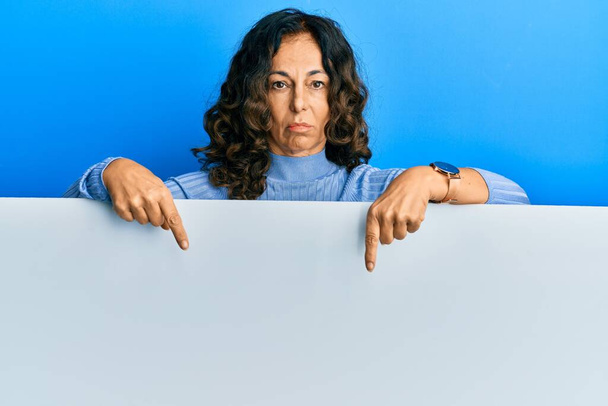 Moyen Age femme hispanique tenant vide bannière pointant vers le bas regardant triste et contrarié, indiquant la direction avec les doigts, malheureux et déprimé.  - Photo, image