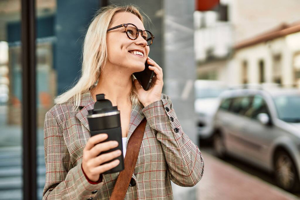 Młoda blondynka bizneswoman rozmawia przez smartfona pijąc kawę w mieście. - Zdjęcie, obraz