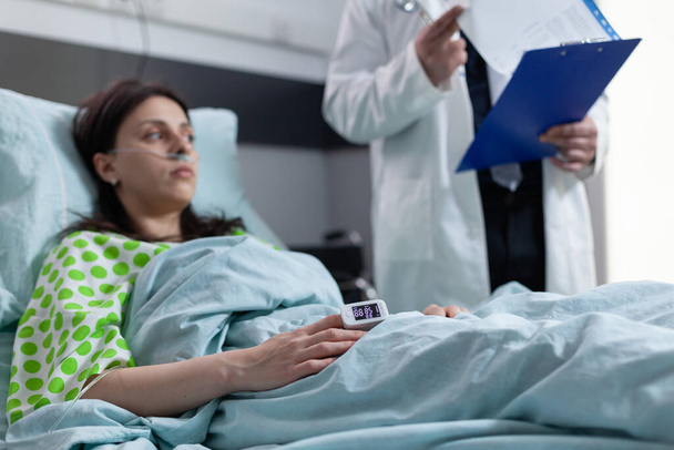 Žena na nemocničním lůžku s pulzním oxymetrem na prstu ukazuje nízkou saturaci kyslíkem poslech lékaře - Fotografie, Obrázek