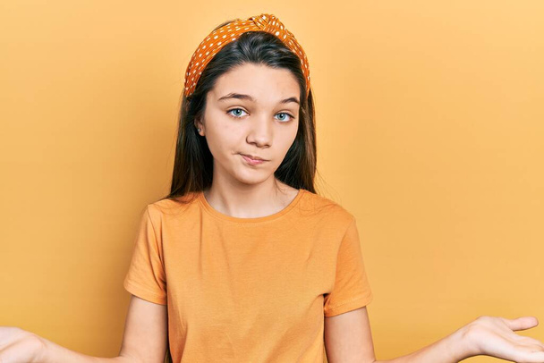Fiatal barna lány visel alkalmi narancssárga póló tanácstalan és zavaros kifejezés a karok és a kezek emelt. kétség.  - Fotó, kép