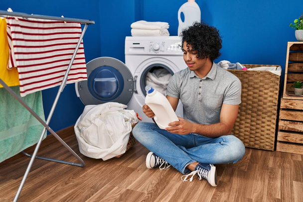 Jonge Spaanse man glimlacht vol vertrouwen terwijl hij flessendetergent vasthoudt in de wasruimte - Foto, afbeelding