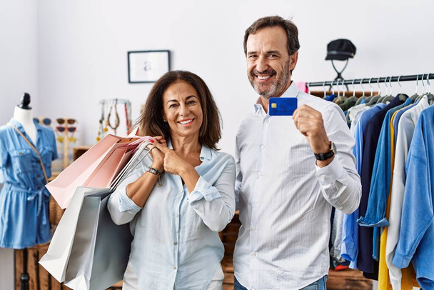 Hispanic middle age couple holding shopping bags and credit card cheerful with a smile on face pointing with hand and finger up to the side with happy and natural expression  - Fotografie, Obrázek