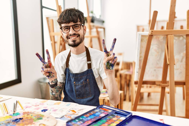 Латиноамериканець з бородою в художній студії посміхається дивлячись на камеру, яка показує пальці, що роблять знак перемоги. номер два.  - Фото, зображення