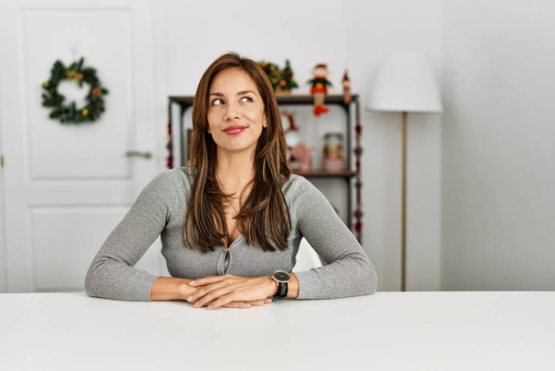 Mujer latina joven sentada en la mesa por la decoración de Navidad sonriendo mirando hacia un lado y mirando hacia otro pensando.  - Foto, Imagen
