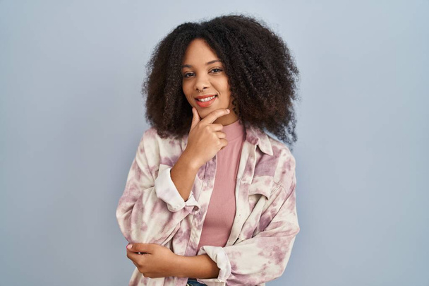 Young african american woman standing over blue background looking confident at the camera smiling with crossed arms and hand raised on chin. thinking positive.  - Foto, afbeelding