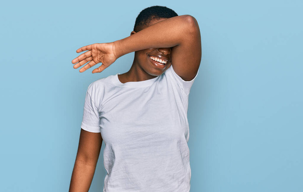 Jonge Afro-Amerikaanse vrouw met casual wit t-shirt over de ogen met een glimlachende arm vrolijk en grappig. blind concept.  - Foto, afbeelding