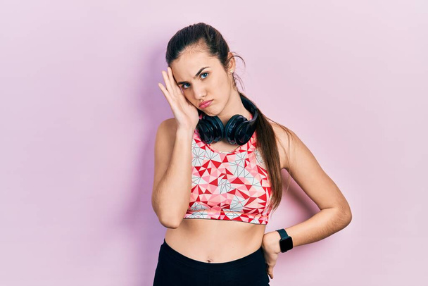 Young brunette teenager wearing gym clothes and using headphones thinking looking tired and bored with depression problems with crossed arms.  - Photo, Image