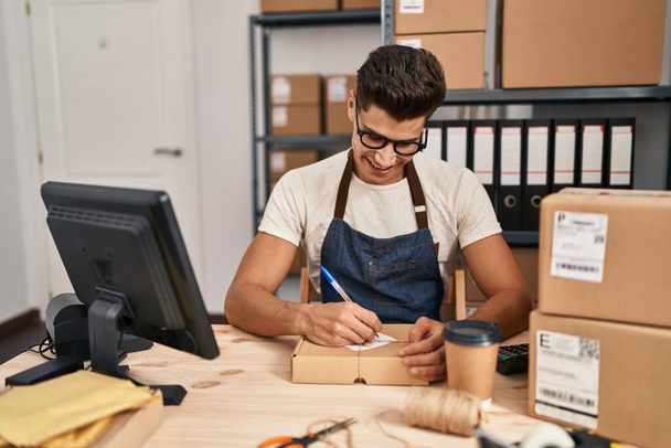Young hispanic man ecommerce business worker writing on package at office - 写真・画像