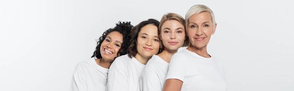 Mujeres multiétnicas en camisetas blancas mirando a la cámara aislada en gris, pancarta  - Foto, imagen
