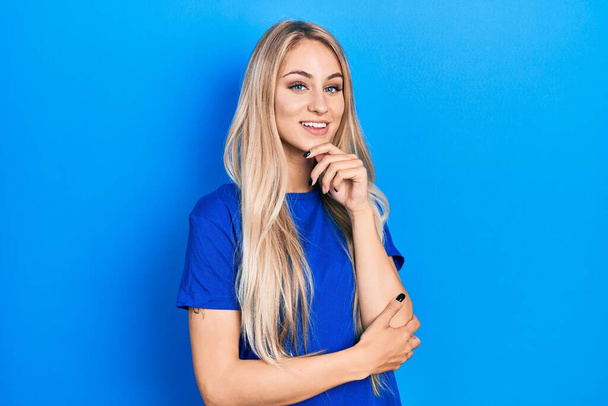 Fiatal, gyönyörű kaukázusi nő alkalmi kék pólóban mosolyog, magabiztosan néz a kamerába keresztbe tett karokkal és kézzel az állán. pozitív gondolkodás.  - Fotó, kép