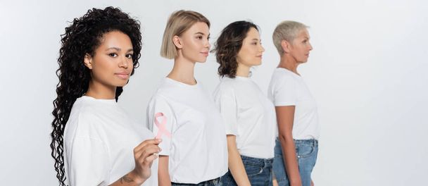 Young african american woman holding pink ribbon of breast cancer awareness near friends isolated on grey, banner  - Zdjęcie, obraz