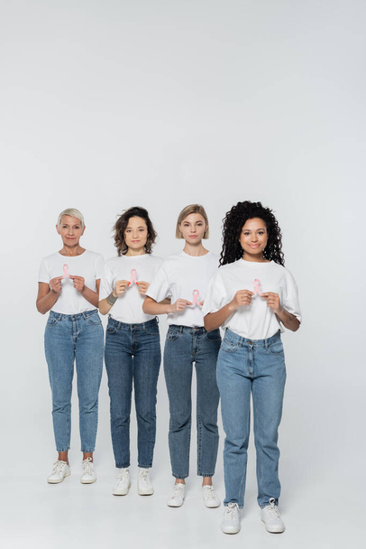 Full length of multiethnic women holding ribbons of breast cancer awareness on grey background  - Foto, afbeelding