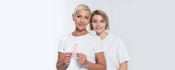 Mujer joven de pie cerca de amigo mayor con cinta de la conciencia del cáncer de mama aislado en gris, pancarta  - Foto, Imagen
