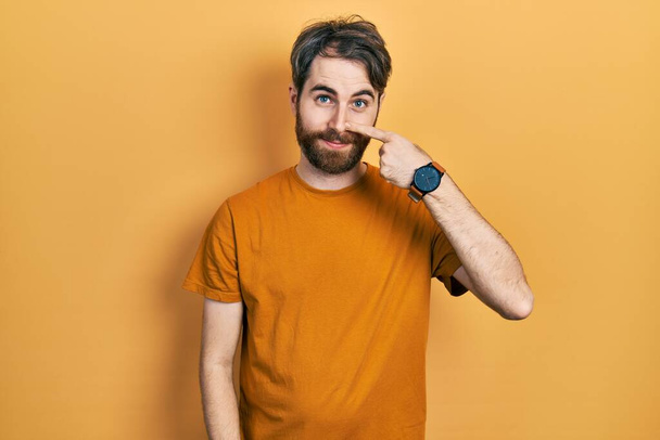 Blanke man met baard in casual geel t-shirt wijzend met de hand vinger naar gezicht en neus, vrolijk glimlachend. schoonheidsconcept  - Foto, afbeelding