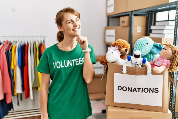 Beautiful caucasian woman wearing volunteer t shirt at donations stand with hand on chin thinking about question, pensive expression. smiling and thoughtful face. doubt concept.  - Photo, Image