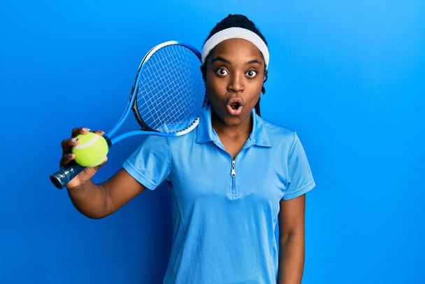 Africká Američanka s copánky hrající tenis držící raketu a míč vystrašený a ohromený s otevřenými ústy pro překvapení, nevěřící tvář  - Fotografie, Obrázek