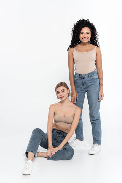 Cheerful african american woman in jeans standing near friend on grey background, feminism concept - Foto, Imagen
