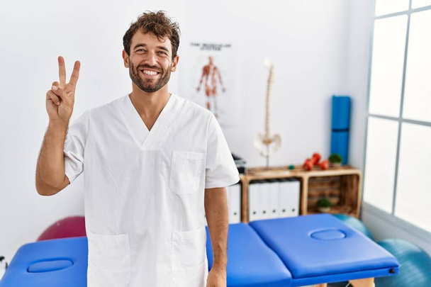 Νεαρός όμορφος φυσιοθεραπευτής που εργάζεται στην κλινική αποκατάστασης πόνου που δείχνει και δείχνει με τα δάχτυλα νούμερο δύο ενώ χαμογελά σίγουρος και χαρούμενος.  - Φωτογραφία, εικόνα