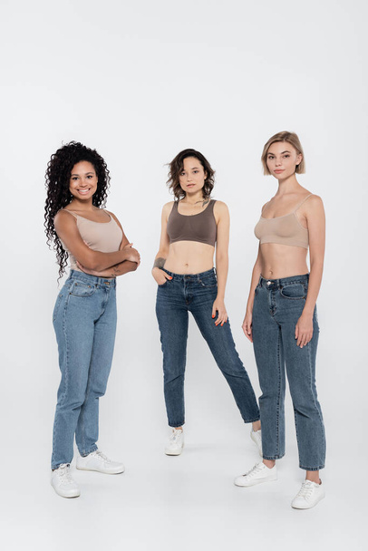 Young interracial women in tops and jeans posing on grey background  - Фото, изображение