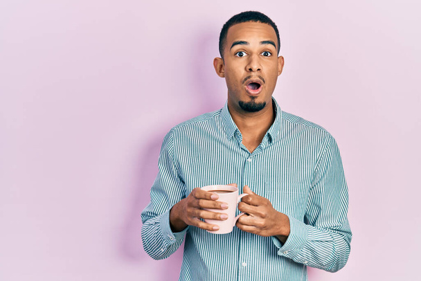 Giovane uomo afroamericano bere una tazza di caffè spaventato e scioccato con sorpresa e stupito espressione, paura e viso eccitato.  - Foto, immagini