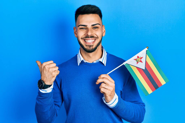 Jonge Spaanse man met baard houdt zimbabwe vlag wijzen duim naar de zijkant glimlachend blij met open mond  - Foto, afbeelding