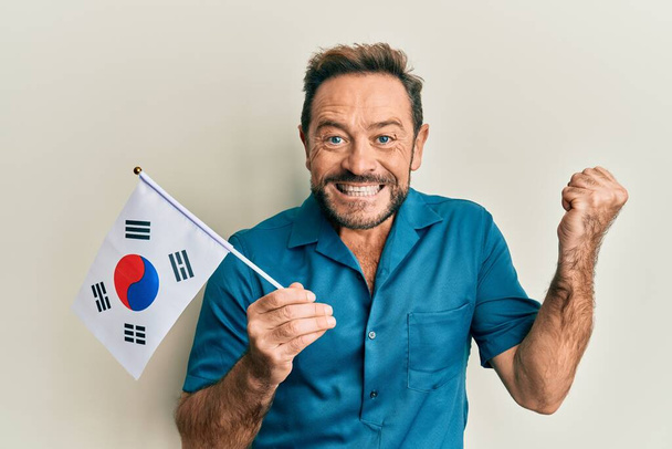 Μεσήλικας άντρας που κρατάει τη σημαία της Νότιας Κορέας ουρλιάζοντας περήφανος, γιορτάζοντας τη νίκη και την επιτυχία πολύ ενθουσιασμένος με σηκωμένο χέρι  - Φωτογραφία, εικόνα