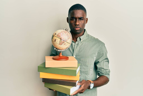 Young african american man holding vintage world ball and books skeptic and nervous, frowning upset because of problem. negative person.  - Foto, imagen