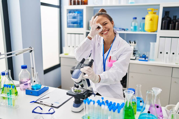 Νεαρή ισπανόφωνη γυναίκα που εργάζεται στο εργαστήριο επιστήμονας χαμογελώντας ευτυχισμένη κάνει εντάξει σημάδι με το χέρι στο μάτι κοιτάζοντας μέσα από τα δάχτυλα  - Φωτογραφία, εικόνα