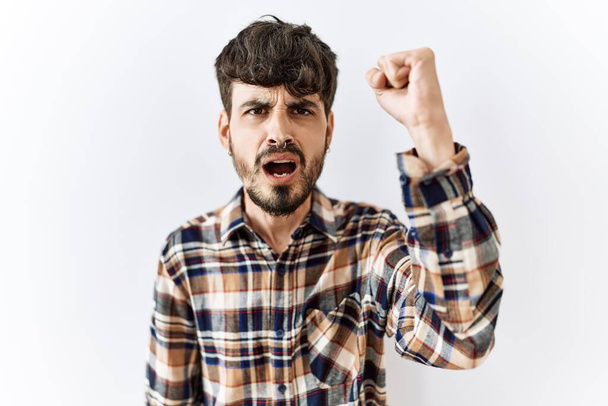 Hombre hispano con barba de pie sobre un fondo aislado enojado y loco levantando el puño frustrado y furioso mientras gritaba con ira. rabia y concepto agresivo.  - Foto, Imagen
