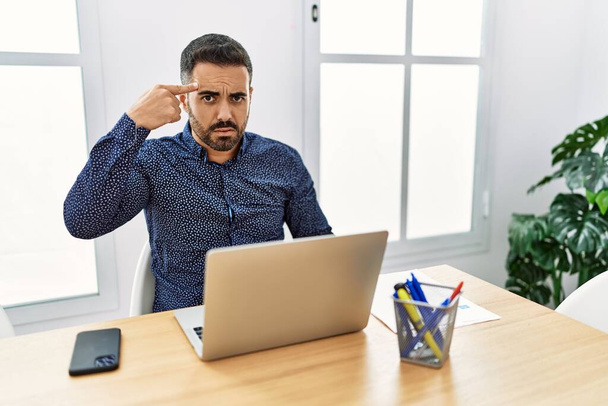 Jeune homme hispanique avec barbe travaillant au bureau avec un ordinateur portable pointant malheureux à boutons sur le front, infection laide de la tête noire. acné et problème de peau  - Photo, image